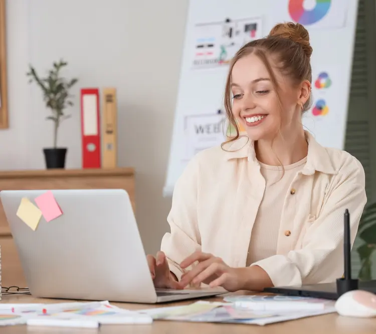 A young woman smiling while working on a laptop at a desk, with colorful charts and sticky notes visible. There are papers, a tablet, and a pen on the desk, and a whiteboard with graphs in the background. -JRPMTS LLC