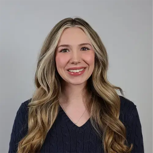 A woman with long, wavy blonde hair smiles at the camera. She is wearing a dark, textured sweater and is posed in front of a plain, light gray background. -JRPMTS LLC