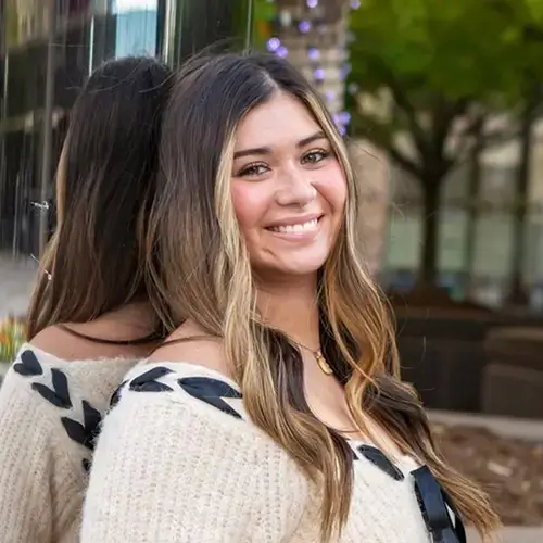 A young woman with long brown hair and blonde highlights smiles while leaning against a reflective glass wall outdoors, with trees and blurred cityscape in the background. -JRPMTS LLC