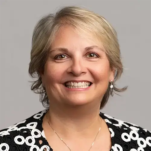 A smiling woman with short, light blonde hair wears a black and white patterned top and small earrings, posed in front of a plain, light gray background. -JRPMTS LLC