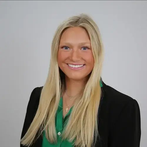 A smiling young woman with long blonde hair wears a black blazer over a green blouse and a gold cross necklace, posing in front of a plain light gray background. -JRPMTS LLC