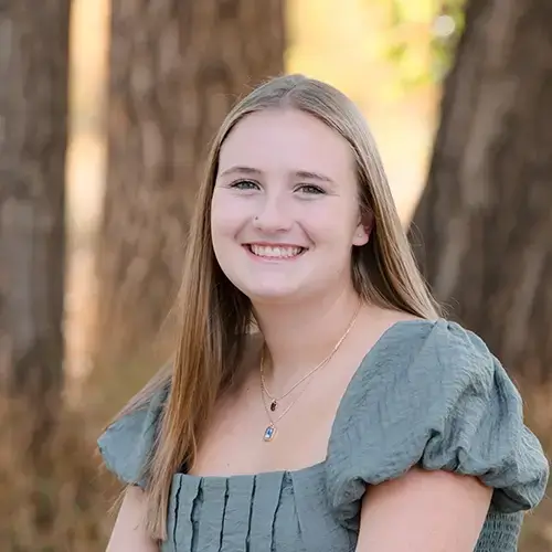 A young woman with long straight blonde hair, wearing a light green short-sleeved dress and a necklace, smiles while sitting outdoors in front of tall trees. -JRPMTS LLC