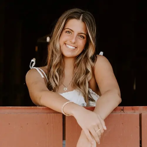 A young woman with long, wavy light brown hair smiles while leaning over a red wooden fence, wearing a white sleeveless top and jewelry, against a dark background. -JRPMTS LLC