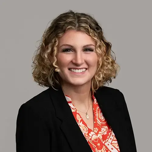 A woman with curly blonde hair, wearing a black blazer over a red and white patterned blouse, smiles at the camera against a plain gray background. -JRPMTS LLC