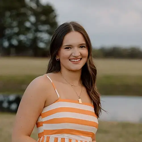 A young woman with long brown hair, wearing a sleeveless orange and white striped dress, stands outdoors in front of a grassy field, trees, and a small pond, smiling at the camera. -JRPMTS LLC