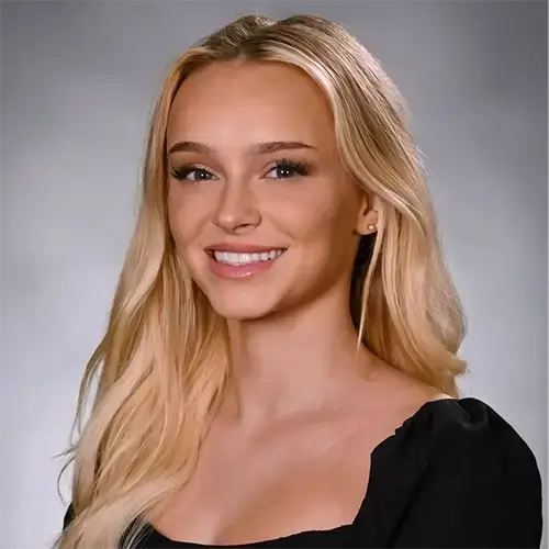 A young woman with long blonde hair, wearing a black top, smiles at the camera in front of a neutral gray background. -JRPMTS LLC