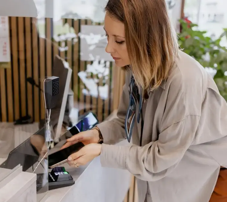 A woman wearing a light-colored shirt uses her smartphone to make a contactless payment at a counter, holding her phone near a card reader inside a modern, well-lit setting. -JRPMTS LLC