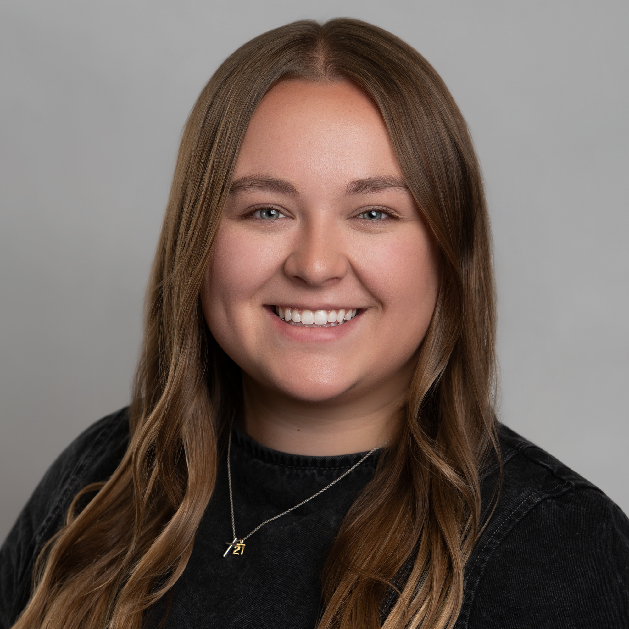 Headshot of a smiling young woman with long brown hair wearing a dark shirt and a silver necklace with a small pendant. -JRPMTS LLC