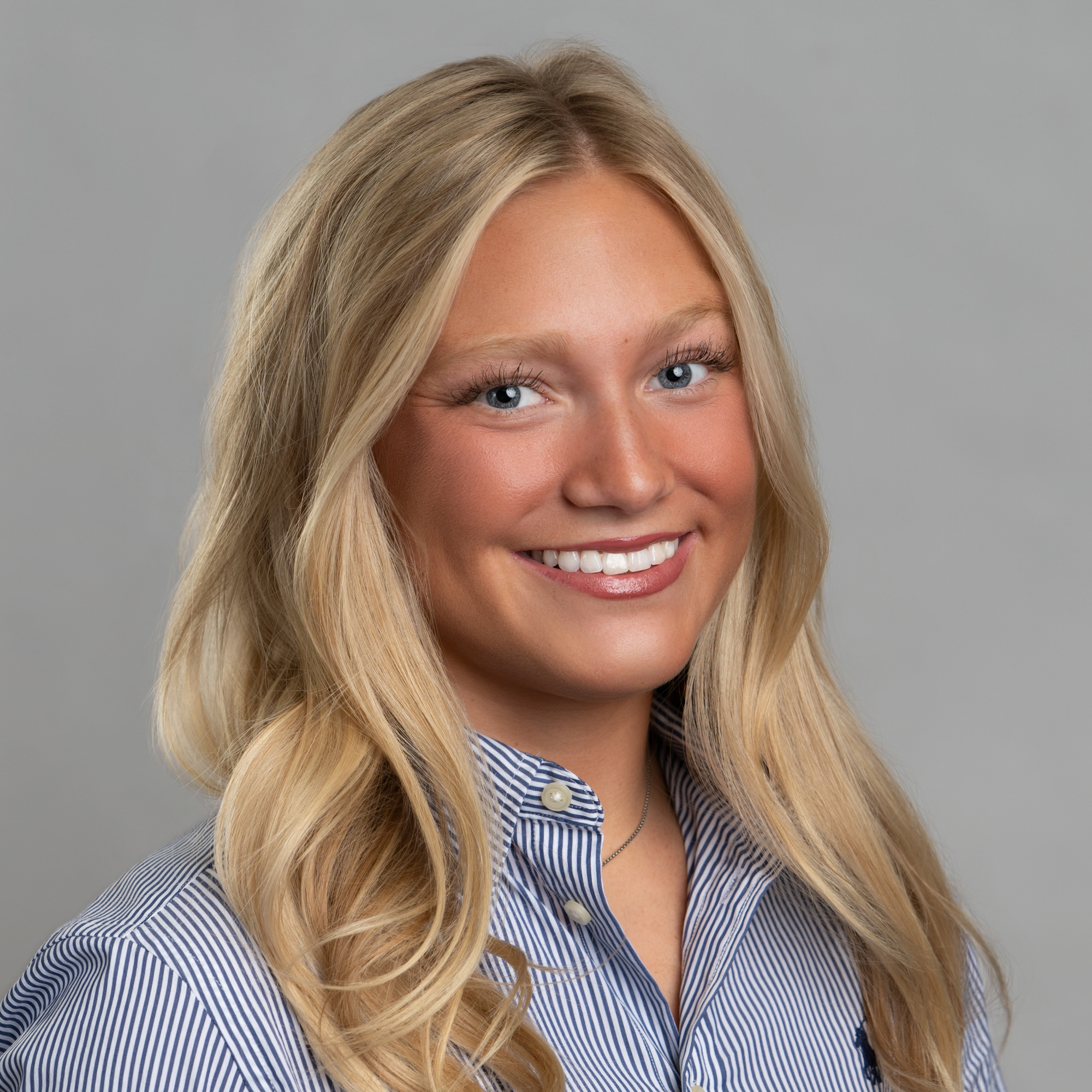 A smiling young woman with long blonde hair wearing a blue and white striped collared shirt. -JRPMTS LLC