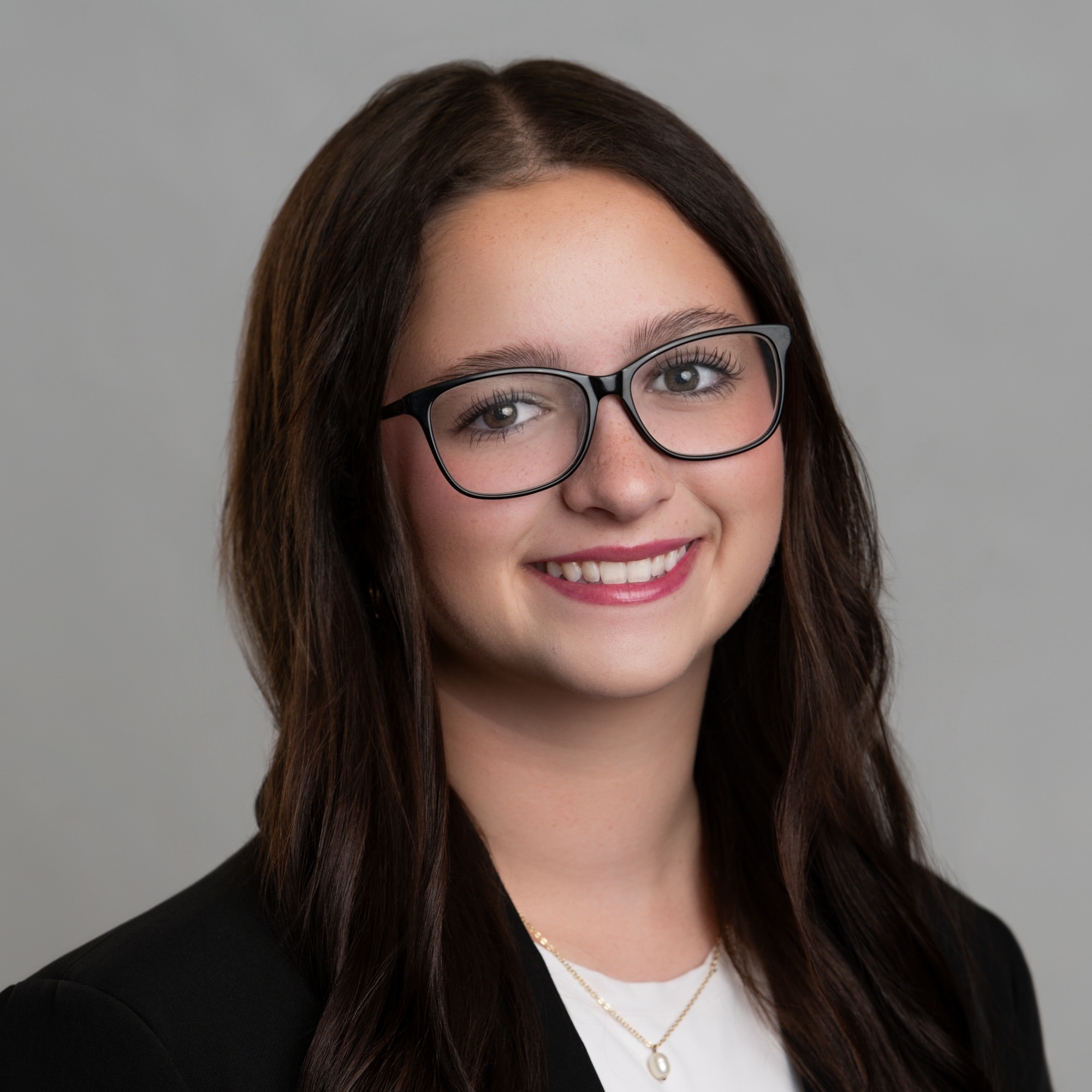 Headshot of a young woman with long dark hair wearing black-rimmed glasses and a black blazer. -JRPMTS LLC
