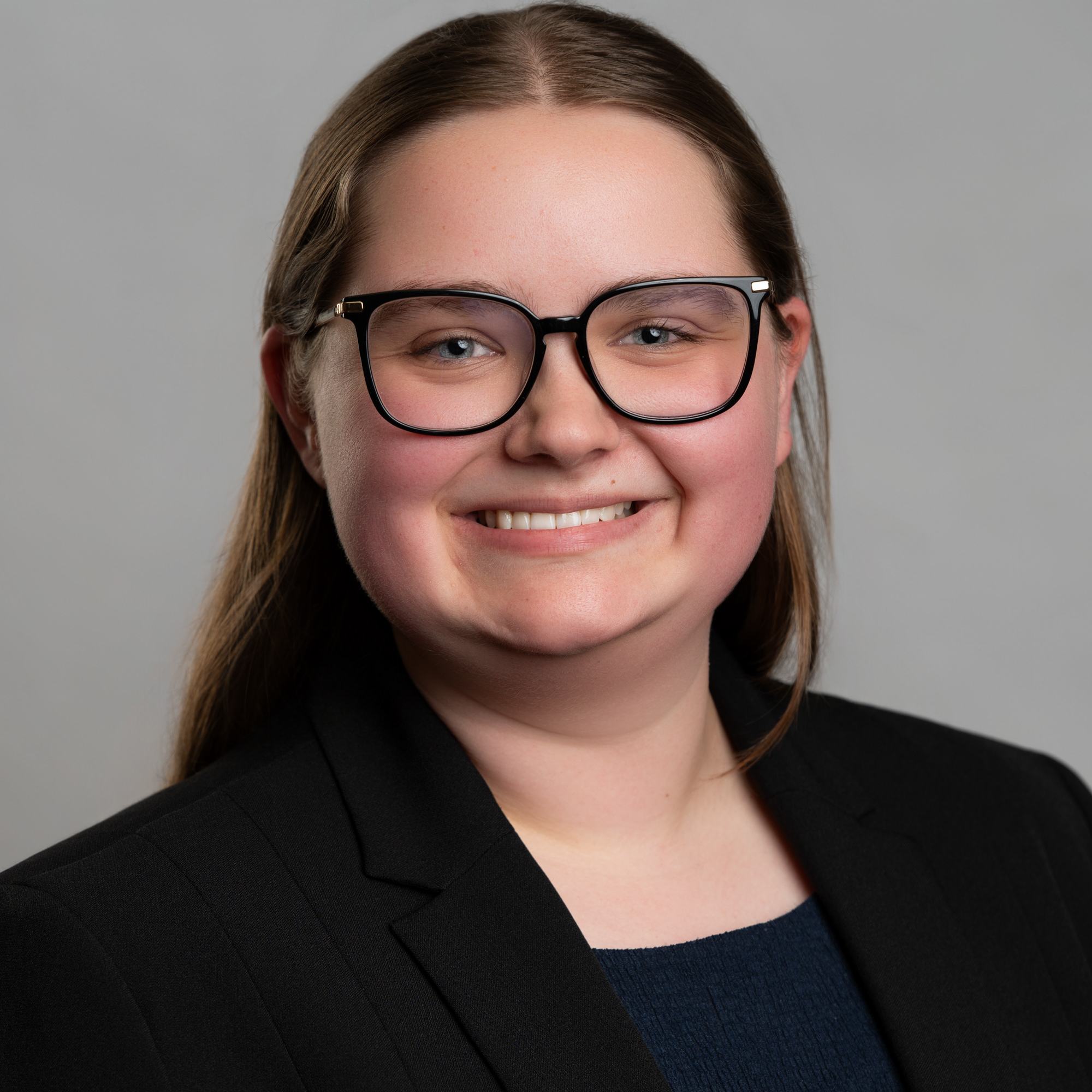 Headshot of a person with long hair and glasses wearing a dark blazer, smiling at the camera. -JRPMTS LLC