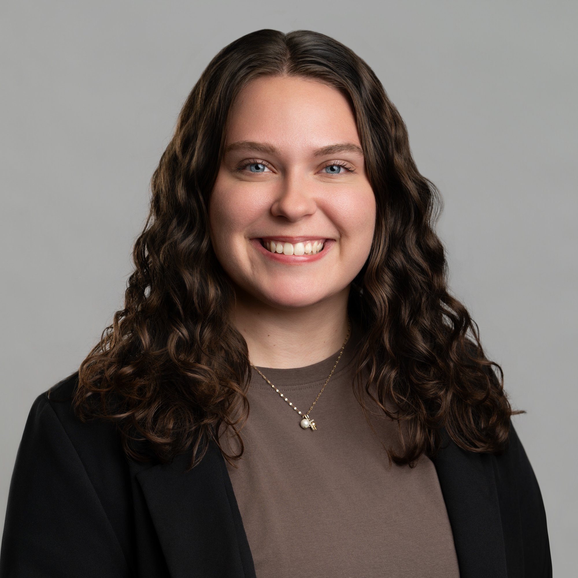 A smiling woman with long wavy hair wearing a brown shirt and a black blazer against a grey background. -JRPMTS LLC